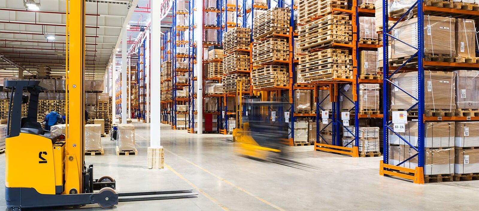 Interior de un centro de distribución amplio y vacío, con muelles de carga, piso de concreto marcado con líneas amarillas, un camión estacionado y dos personas cerca de una puerta abierta.